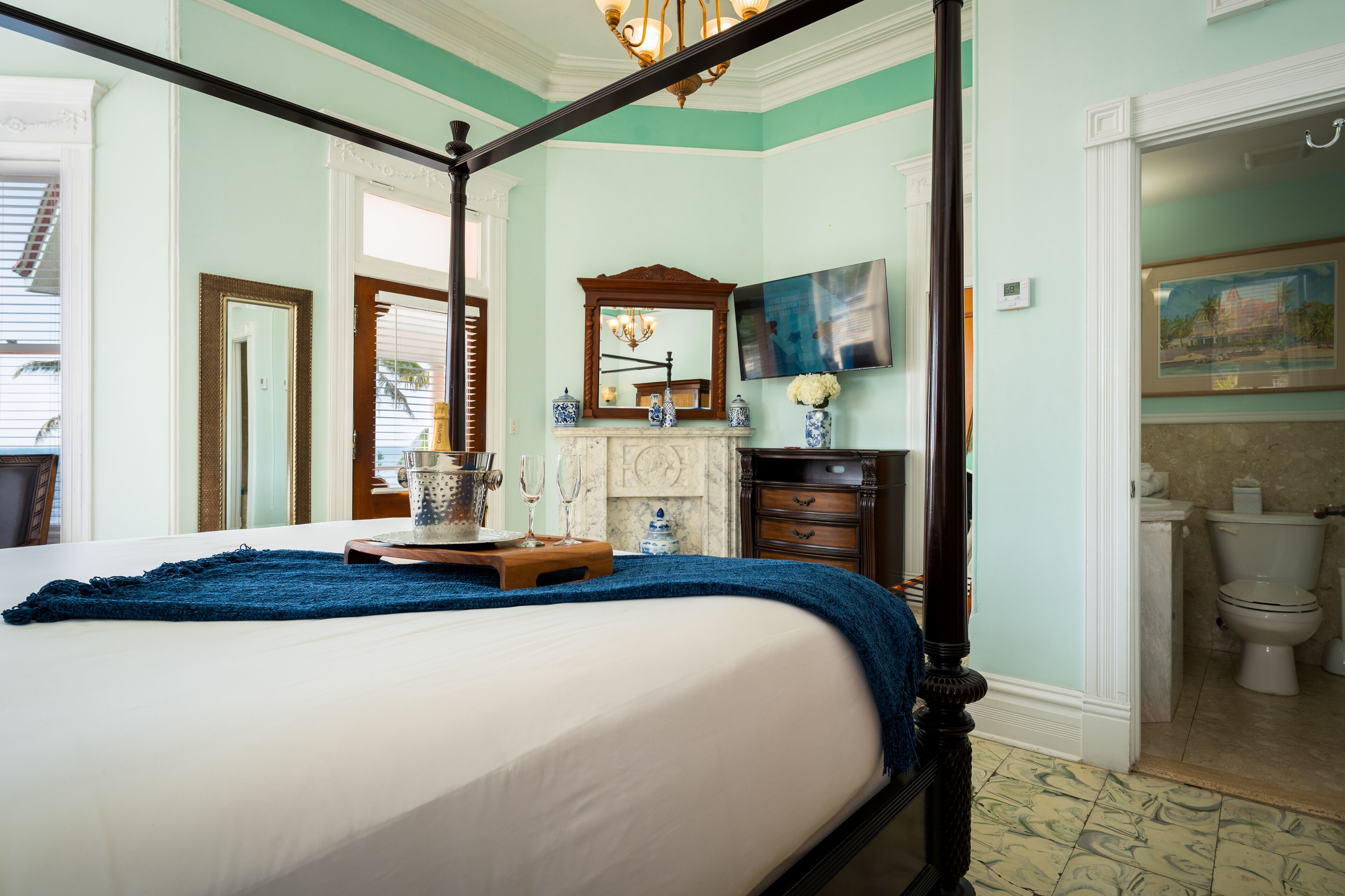 A four-poster bed with a blue throw blanket, a wooden tray with glasses, a mirror, and a painting on the wall.
