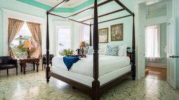 Una cama con dosel de madera, un edredón blanco y una manta azul. Hay dos mesitas de noche con lámparas, una silla y una ventana con cortinas.
