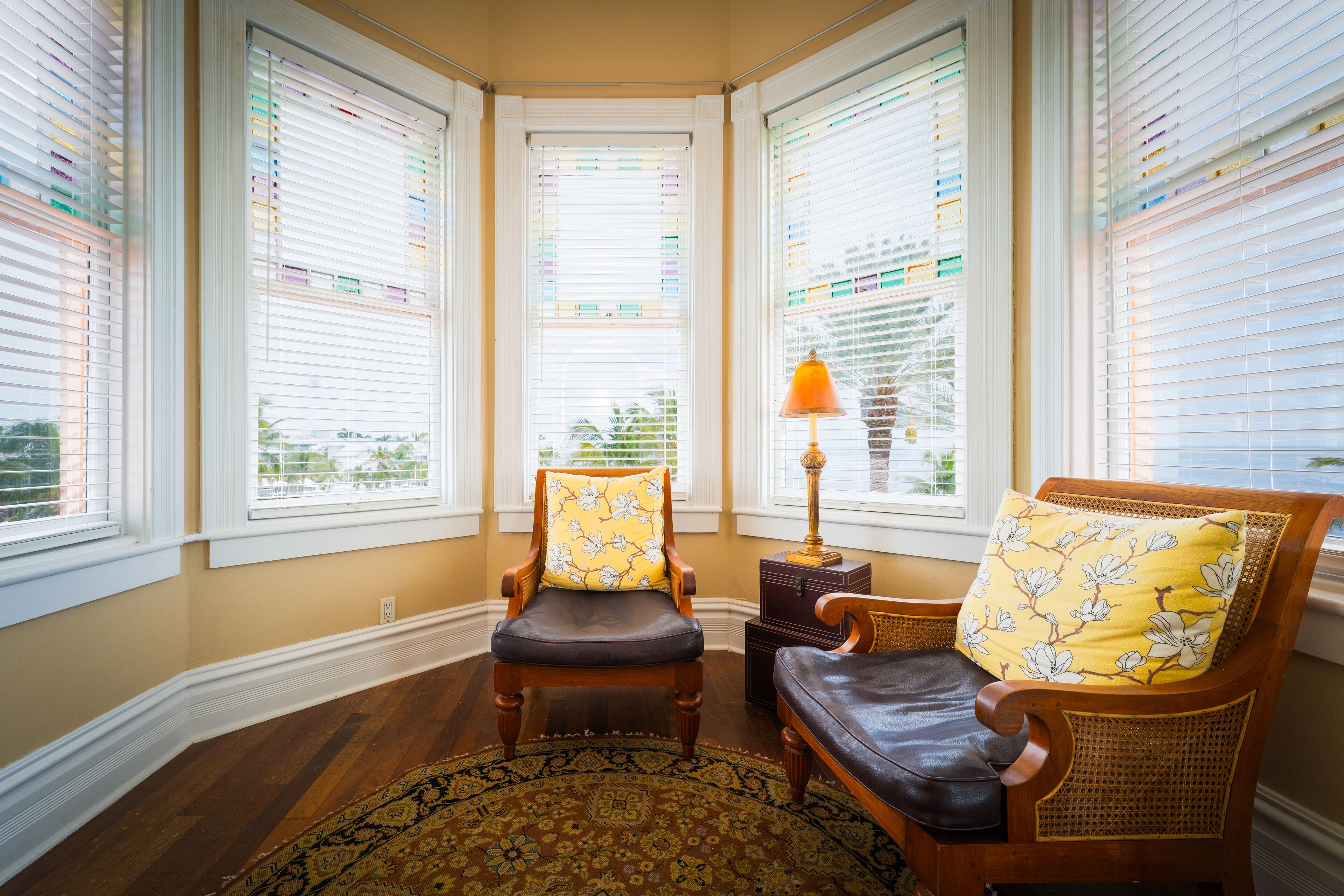 A corner room with a wooden chair and a sofa, both with floral cushions, a small table with a lamp, and a patterned rug.