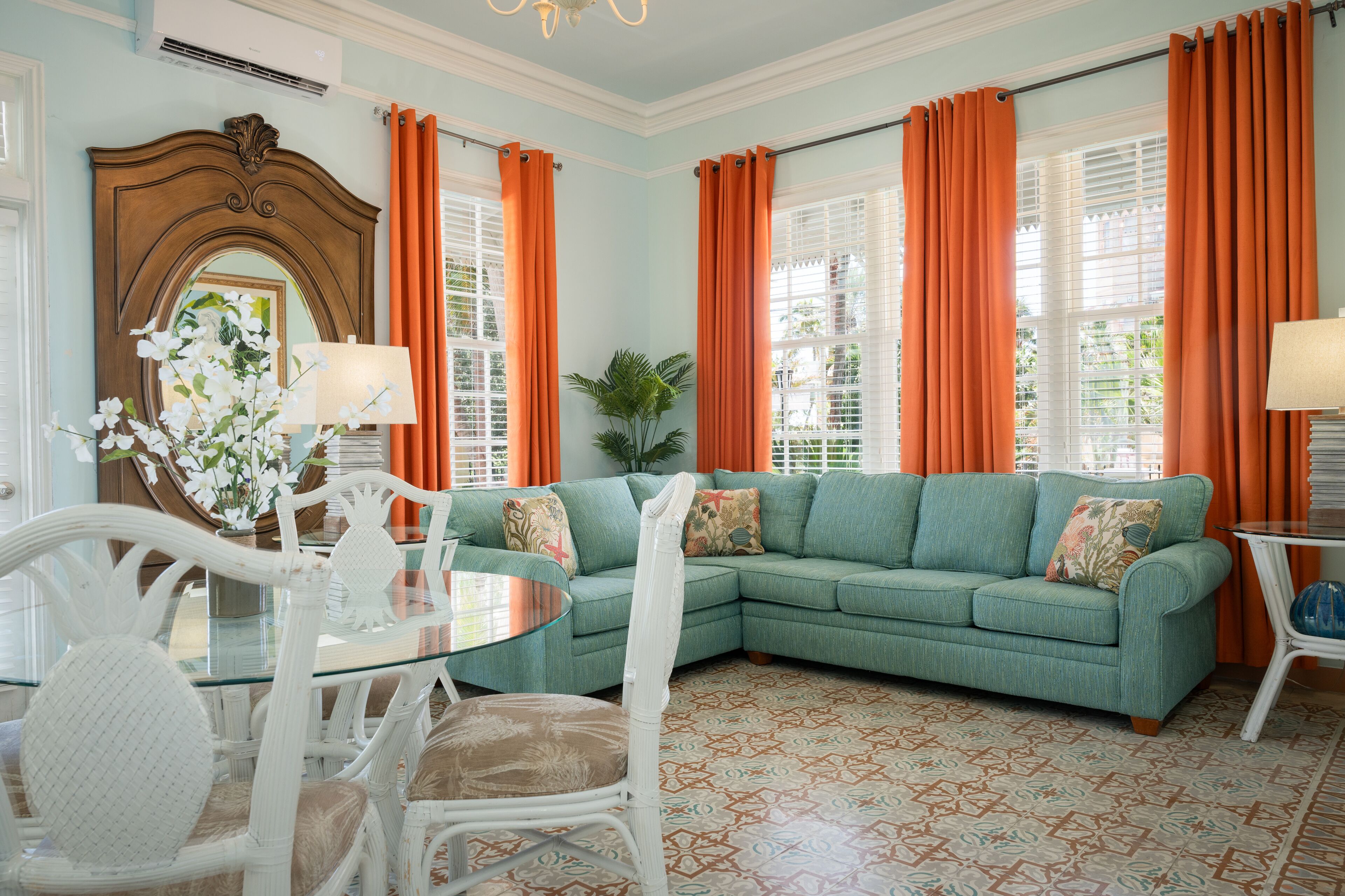 A living room with a green sofa, white chairs, a glass coffee table, and orange curtains.