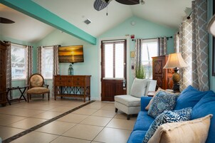 Un salón con un sofá azul, un aparador de madera y una silla. Hay una puerta con ventana y cortinas.