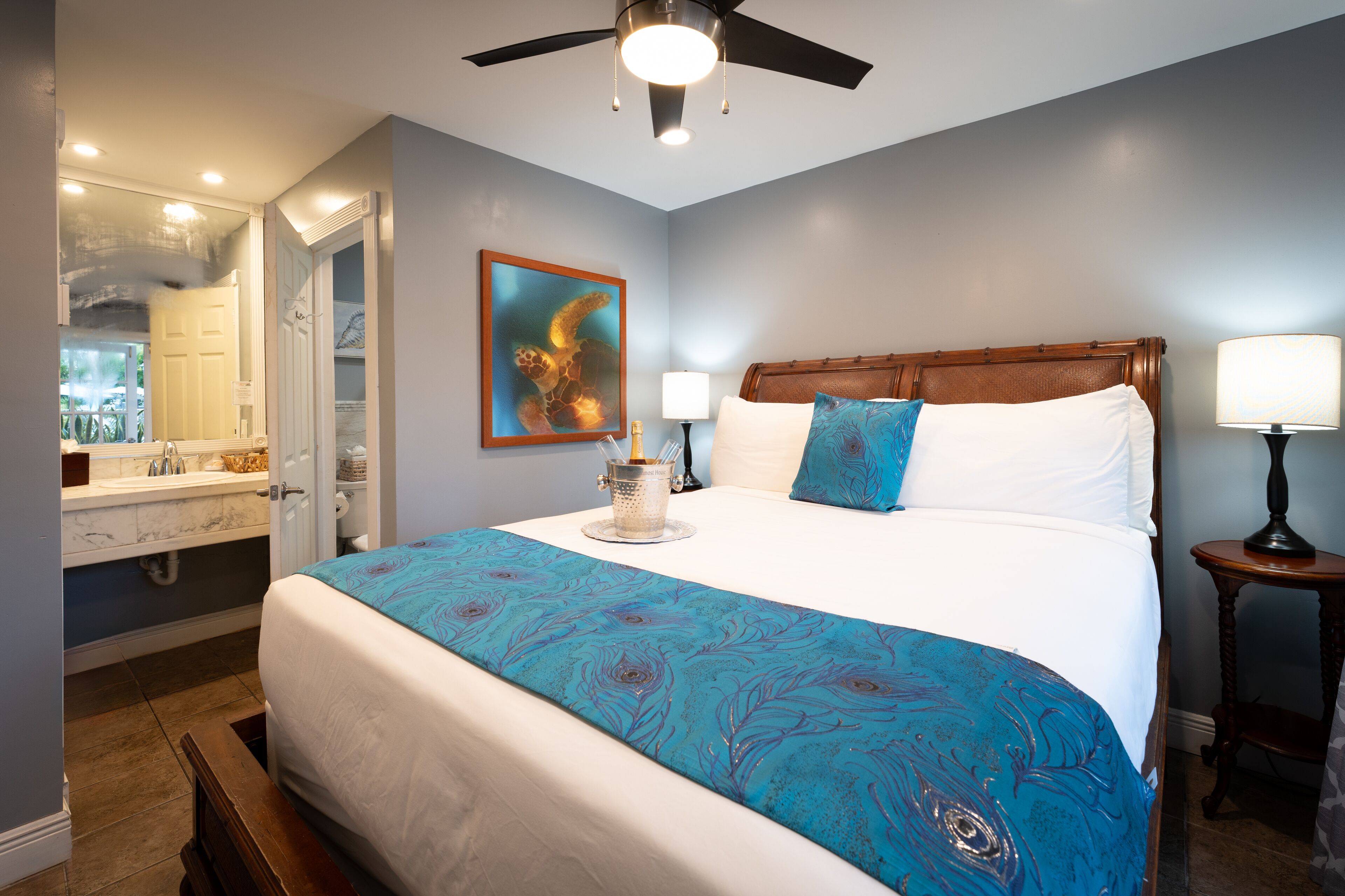 A bedroom with a bed, a ceiling fan, a nightstand, a lamp, a painting, and a bathroom visible through an open door.