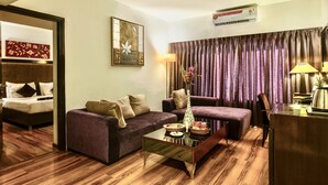 A hotel room with a purple sofa, a wooden coffee table with snacks, a desk with a lamp, and a large window with curtains. - GIS Select Banjara  Hills (Hyderabad)