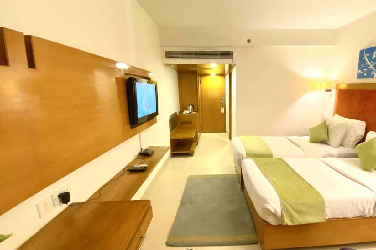 Habitación de hotel con dos camas, un televisor montado en la pared, un escritorio con computadora y un baño visible a través de una puerta abierta.