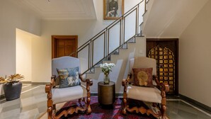 A grand staircase leading to an upper floor, two ornate armchairs with cushions, a vase with flowers, and a patterned rug. - Barwara Kothi (Jaipur)