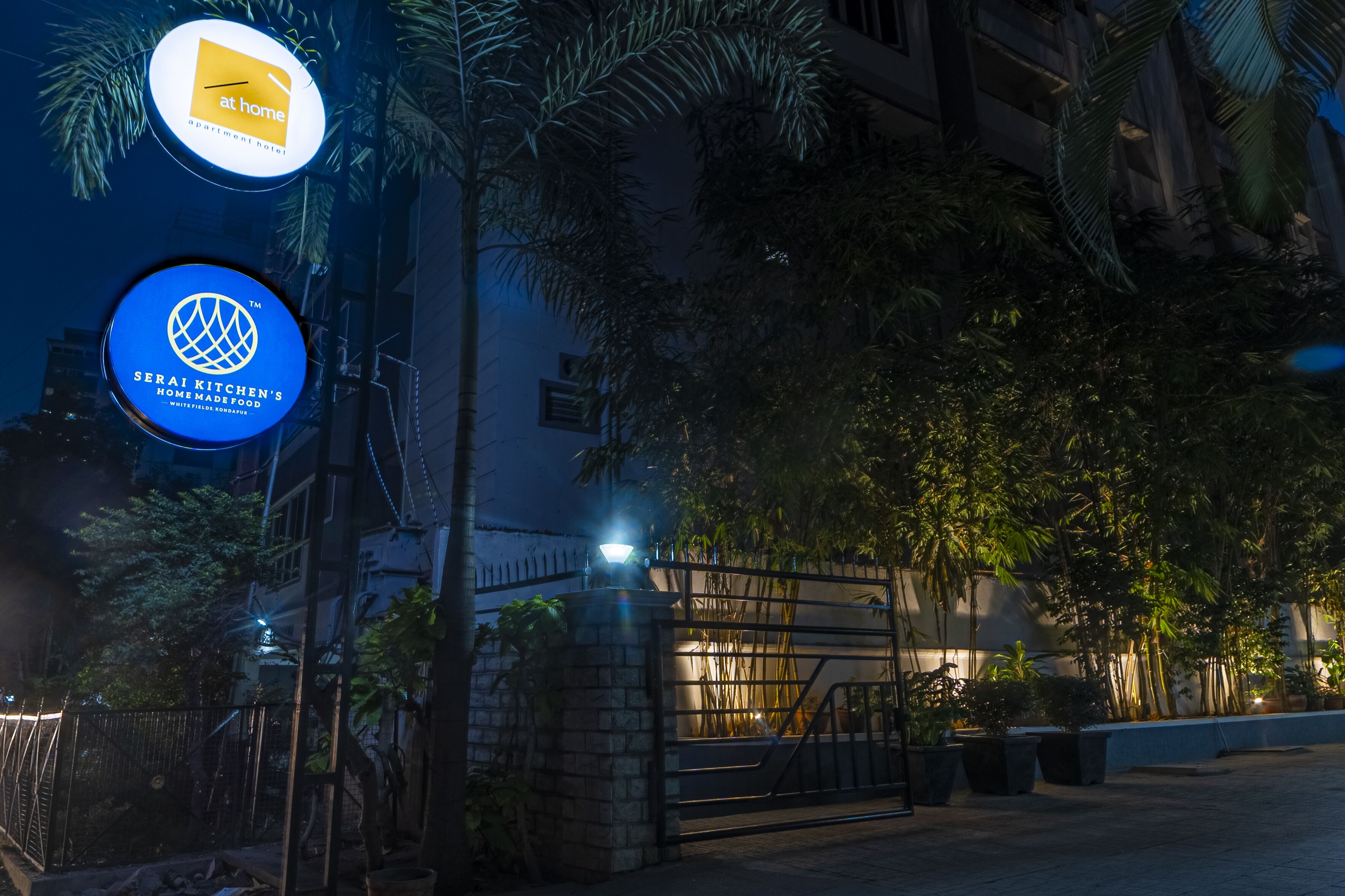 Une entrée sécurisée, agrémentée d’enseignes lumineuses indiquant « ouvert » et « plats faits maison de Serai Kitchen ».