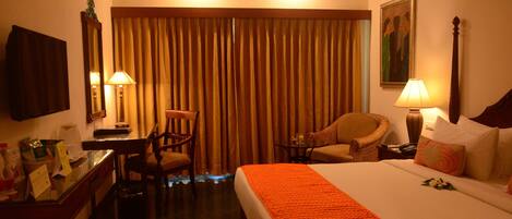 A hotel room with a bed, two chairs, a desk, a lamp, and a painting on the wall.