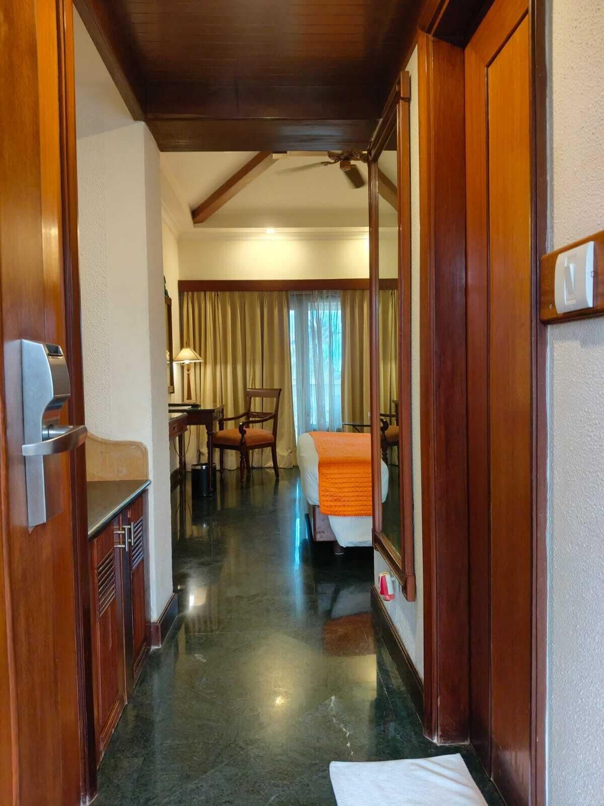 A hotel room with a wooden door, a bed with an orange blanket, a desk with a lamp, and a chair.