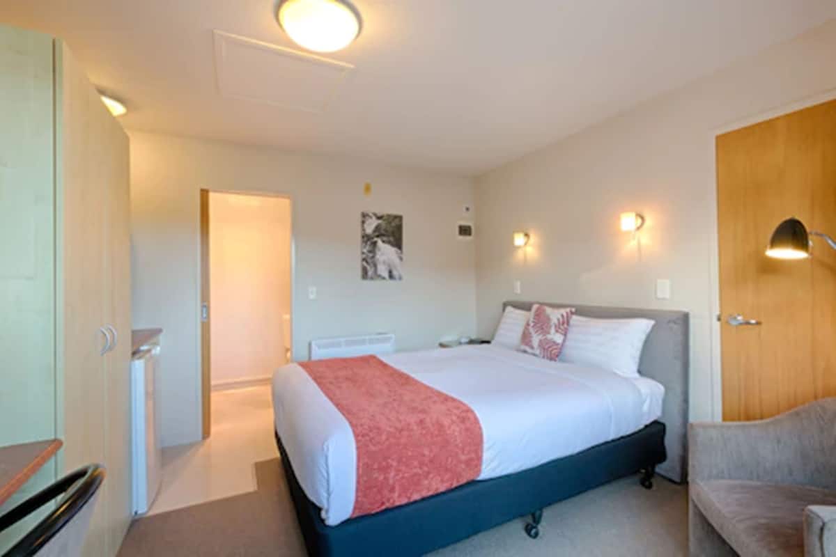 A hotel room with a bed, a chair, a door, and a wall-mounted light fixture.