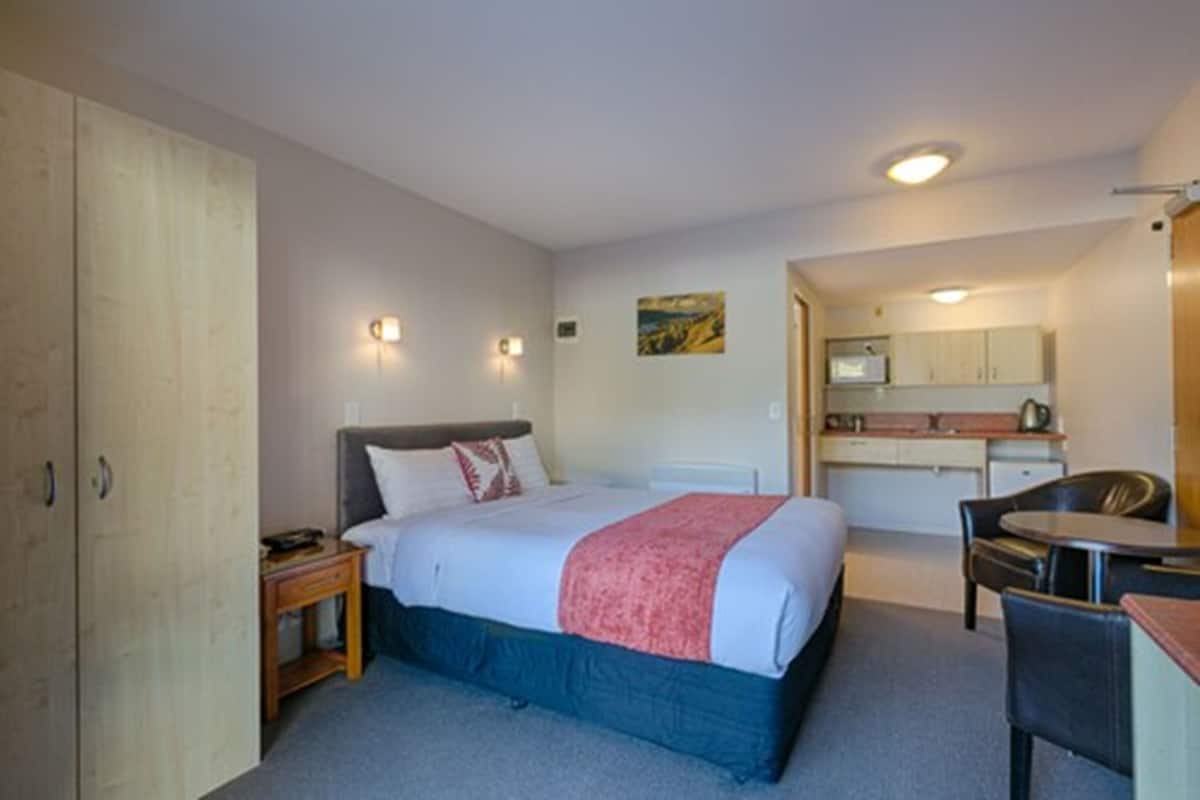 A bedroom with a bed, bedside table, and a small desk with a chair. There is a kitchenette in the background with a microwave and a small refrigerator.