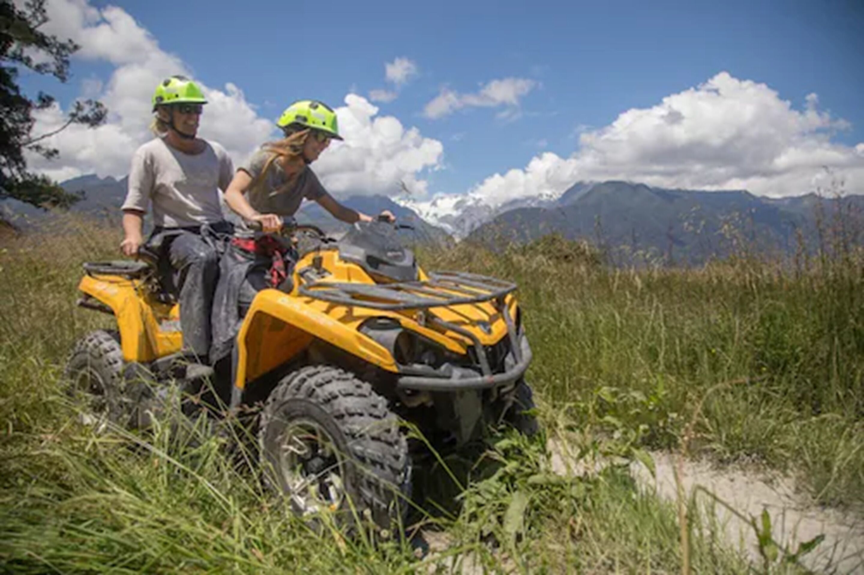 Dos personas en un vehículo todo terreno amarillo sobre una zona herbosa, con montañas al fondo.
