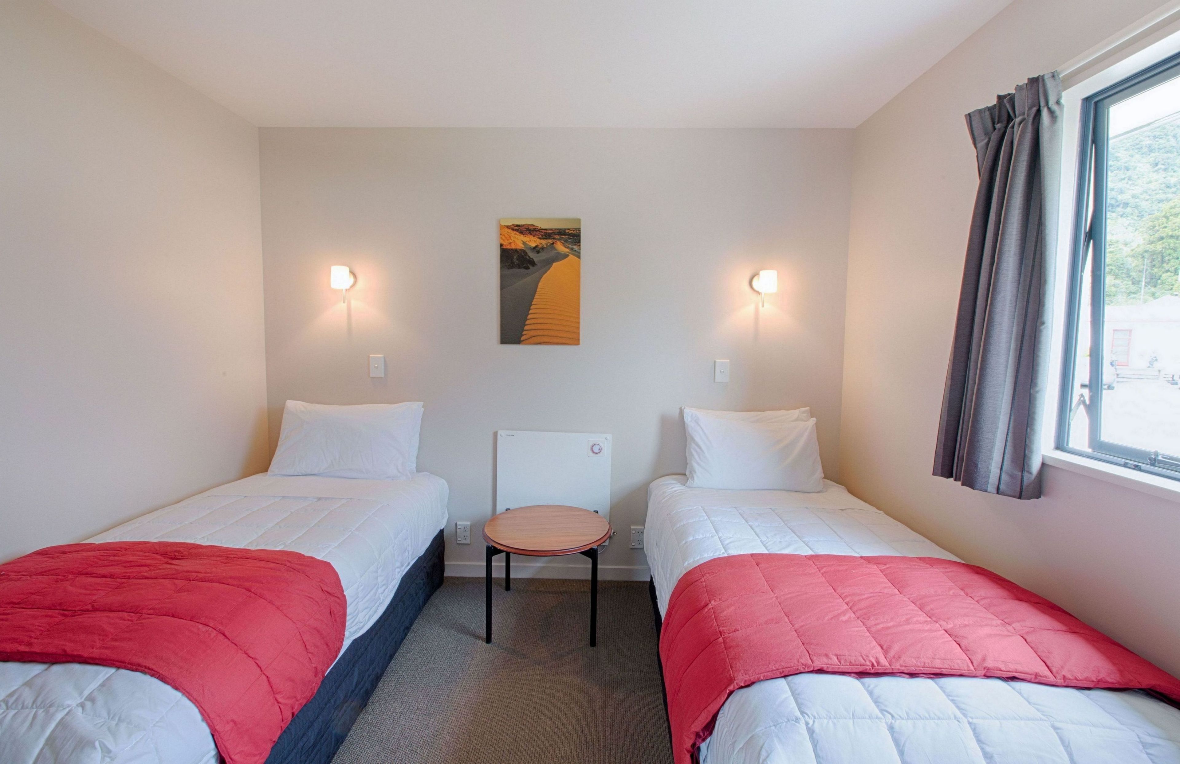 A hotel room with two single beds, a small table, a chair, and a painting on the wall.