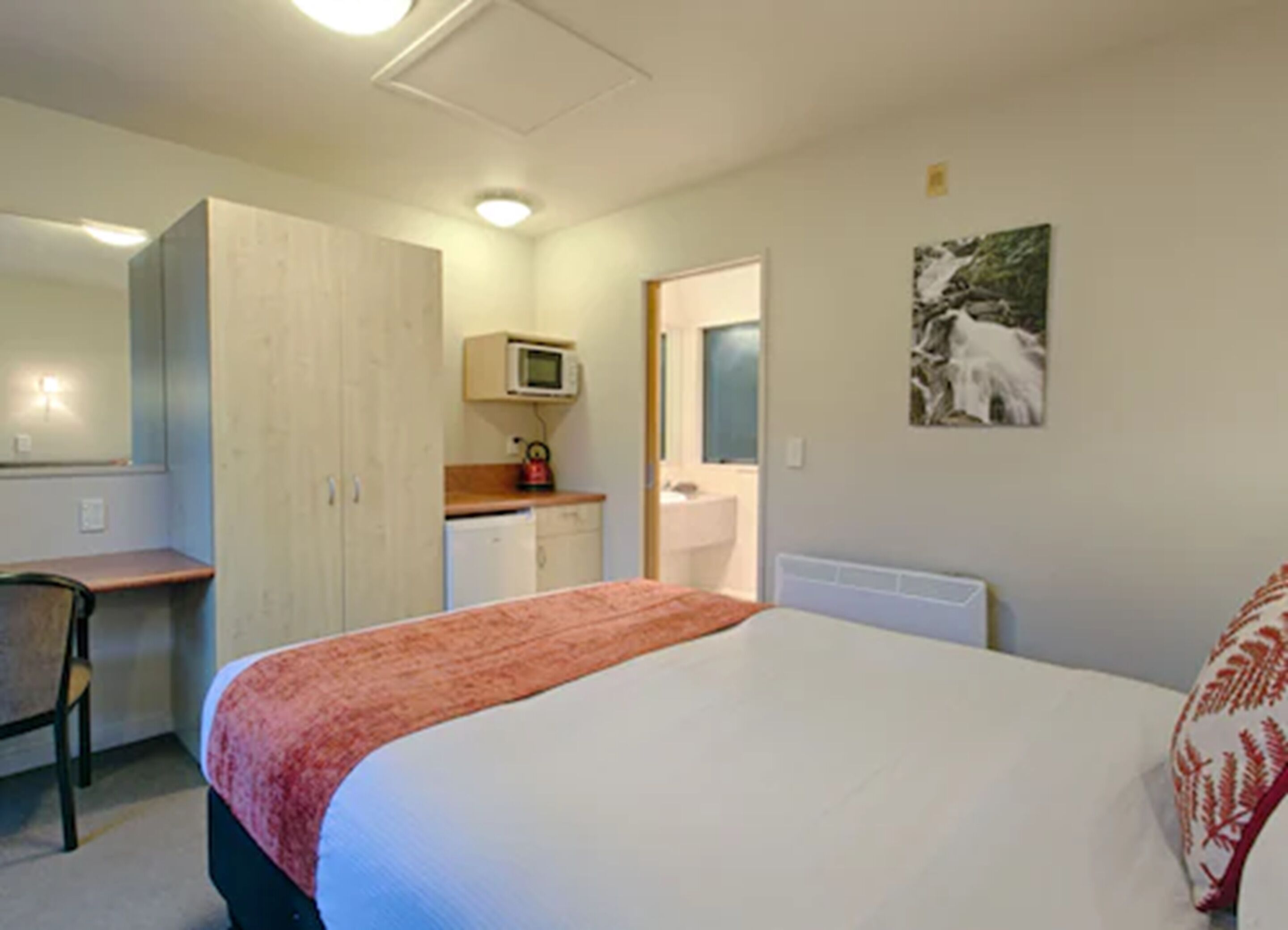 A bedroom with a bed, a desk, a microwave, a sink, and a mirror.