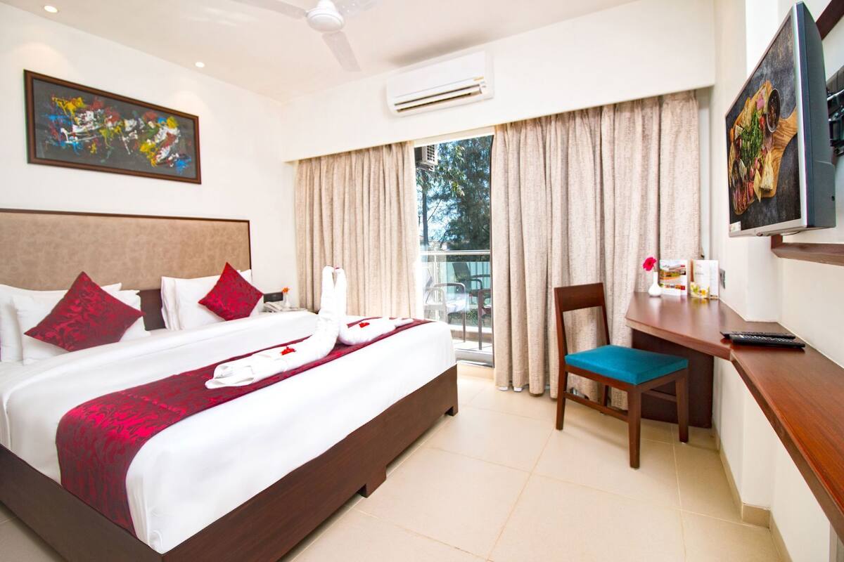 A hotel room with a large bed, a desk with a chair, a TV, and a balcony with curtains.