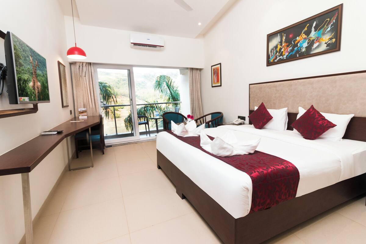 A hotel room with a large bed, a desk, a chair, a balcony with a view of palm trees, and a painting on the wall.