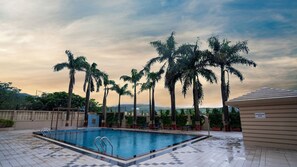 A swimming pool with a tiled deck, surrounded by palm trees and a view of distant hills. - 7 Apple Resort Lonavala (Mawal)