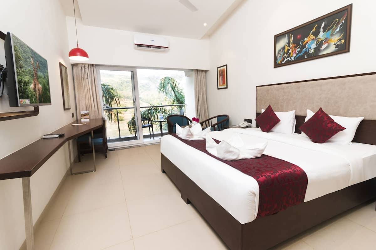 A hotel room with a large bed, a desk, a chair, a balcony with a view of palm trees, and a painting on the wall.