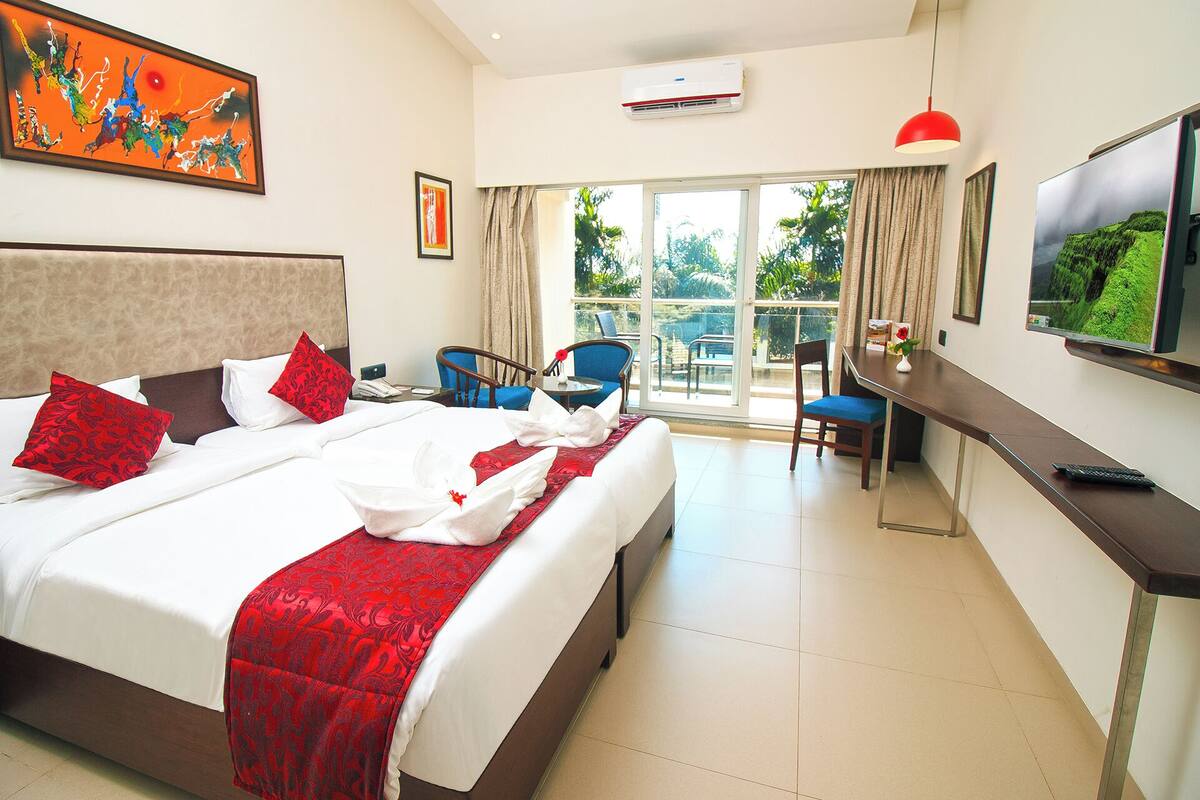 A hotel room with a large bed, a desk with a chair, a TV, and a balcony with curtains.