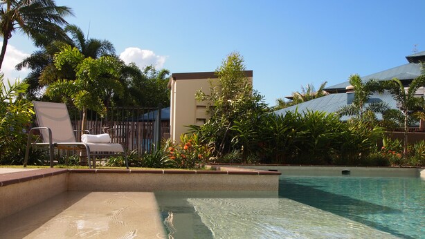 A clear swimming pool with a lounge area, palm trees, and a building in the background. - Edge Apartments Cairns (Cairns North)