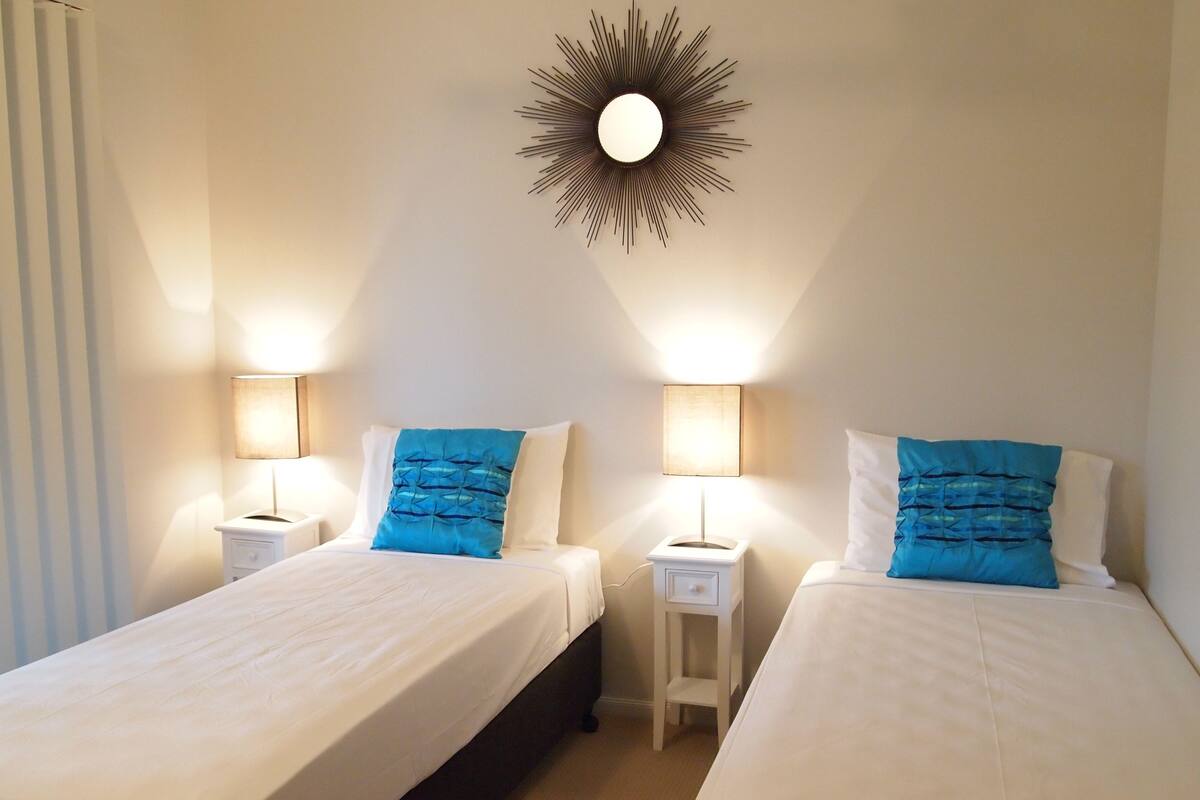 Habitación de hotel con dos camas individuales, cada una con una colcha blanca y almohada azul. Se observan mesas de noche con lámparas, una lámpara de pared y una ventana con persianas.