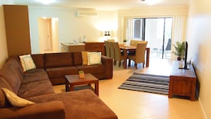 A living room with a brown sofa, a dining table with chairs, and a kitchen area in the background. - Edge Apartments Cairns (Cairns North)