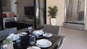 A dining area with a glass table set for four, a television in the background, and a sliding glass door leading to a living room. - Edge Apartments Cairns (Cairns North)