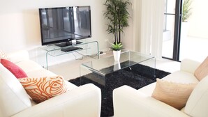 A modern living room with a flat-screen TV on a glass table, white sofas with patterned cushions, a large potted plant, and a sliding glass door leading to an outdoor area. - Edge Apartments Cairns (Cairns North)