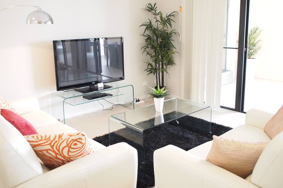 Un salón moderno con un televisor de pantalla plana sobre una mesa de vidrio, sofás blancos con cojines estampados, una planta grande en maceta y un ventanal corredero que da a un espacio exterior.