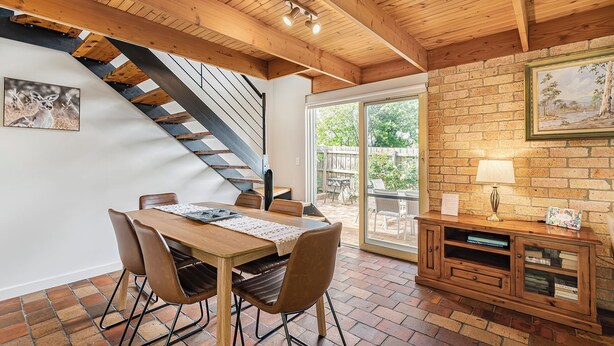 A dining area with a wooden table and chairs, a staircase, and a brick wall with a picture frame. - Bungunyah Apartments (Croydon)