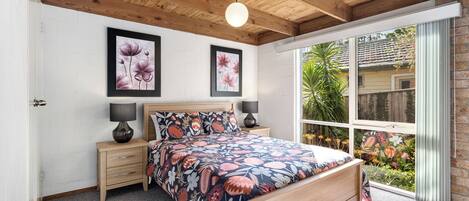 A bedroom with a wooden ceiling, a bed with floral bedding, two bedside lamps, and a large window with a view of a garden.