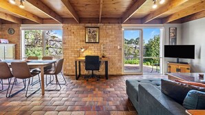 A room with a brick wall, wooden ceiling beams, a dining table with chairs, a sofa, a TV, and a desk with a lamp. - Bungunyah Apartments (Croydon)