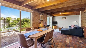 A spacious living area with a dining table, chairs, a sofa, and a TV. There is a large window providing a view of the outdoors. - Bungunyah Apartments (Croydon)