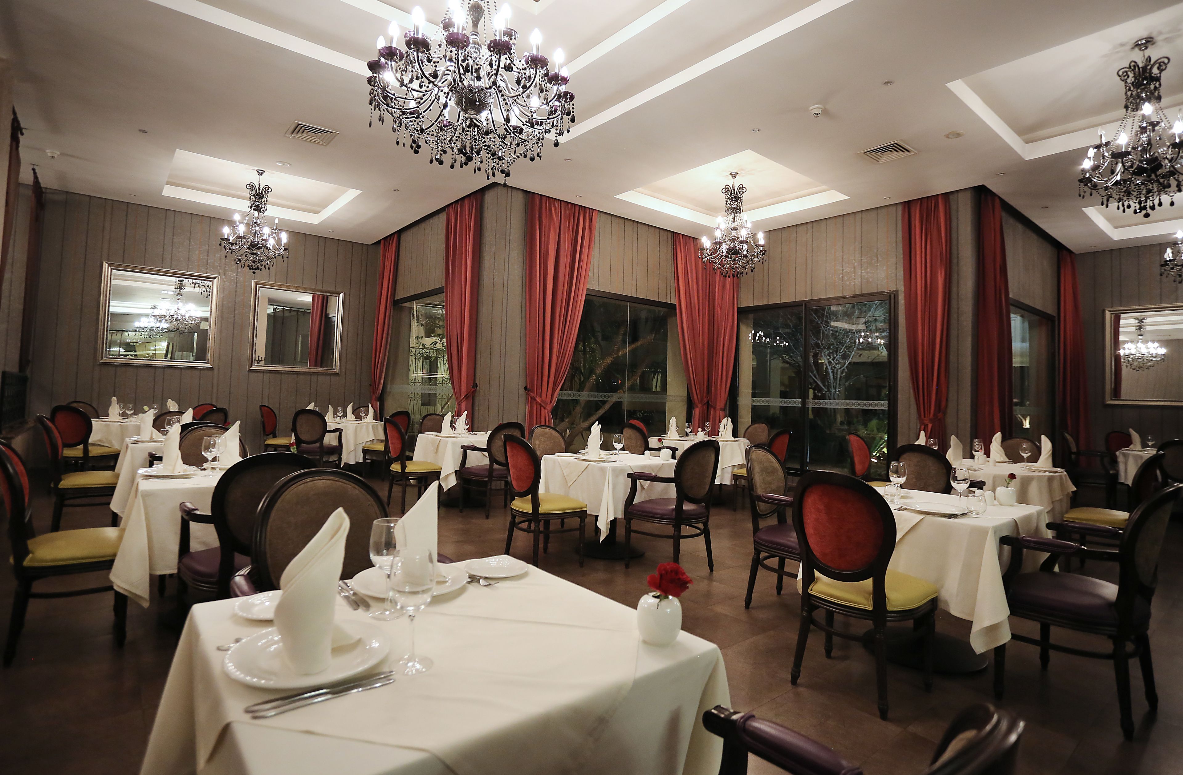 A formal dining room with multiple tables set for service, large windows with red curtains, and ornate chandeliers.