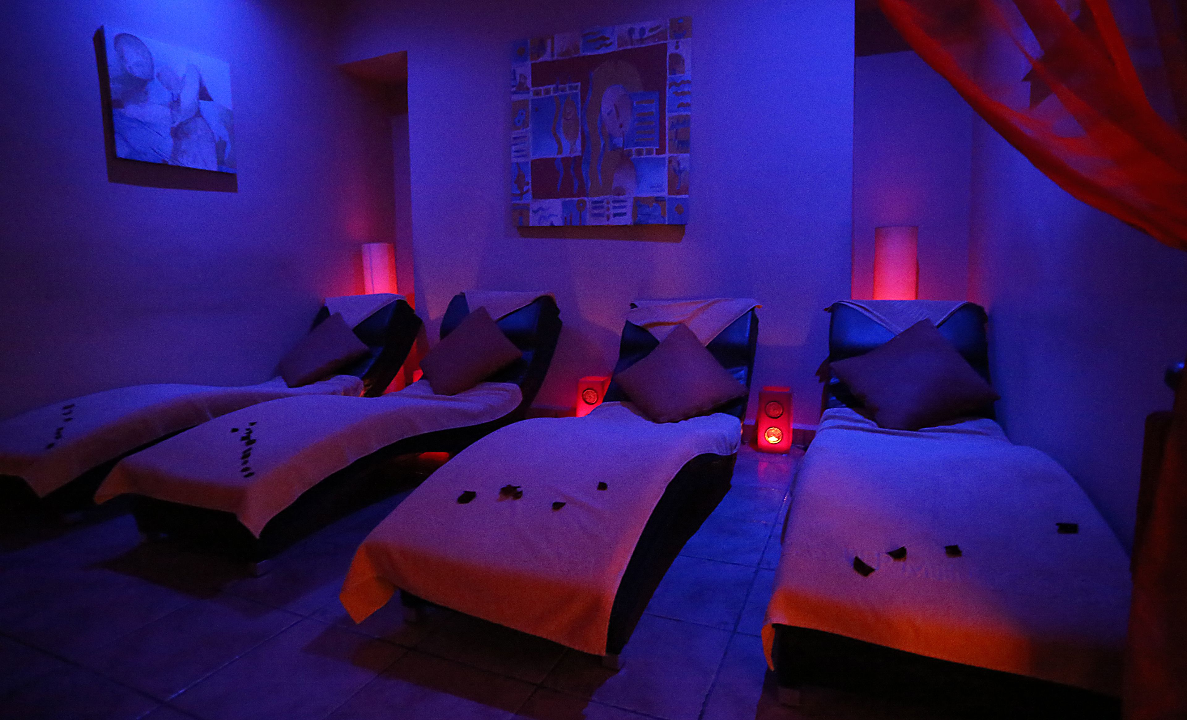 A dimly lit room with three beds, each with a pillow and a blanket. There are wall-mounted lamps and a framed picture above the beds.