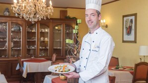 A chef in a formal uniform holding a plate of food in a dining room with a chandelier, wooden cabinets, and a table set for dining. - Hostellerie Val Fleuri (Mersch)