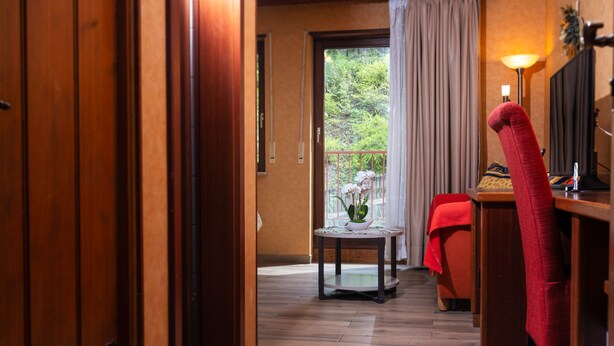 A hotel room with a wooden floor, a small round table with a vase, a red chair, and a bed with a red blanket. - Hostellerie Val Fleuri (Mersch)