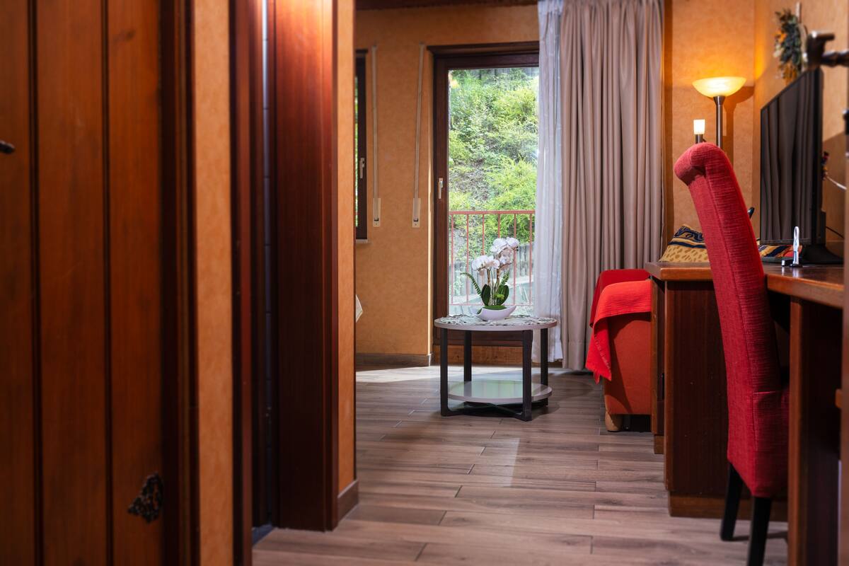 A hotel room with a wooden floor, a small round table with a vase, a red chair, and a bed with a red blanket.