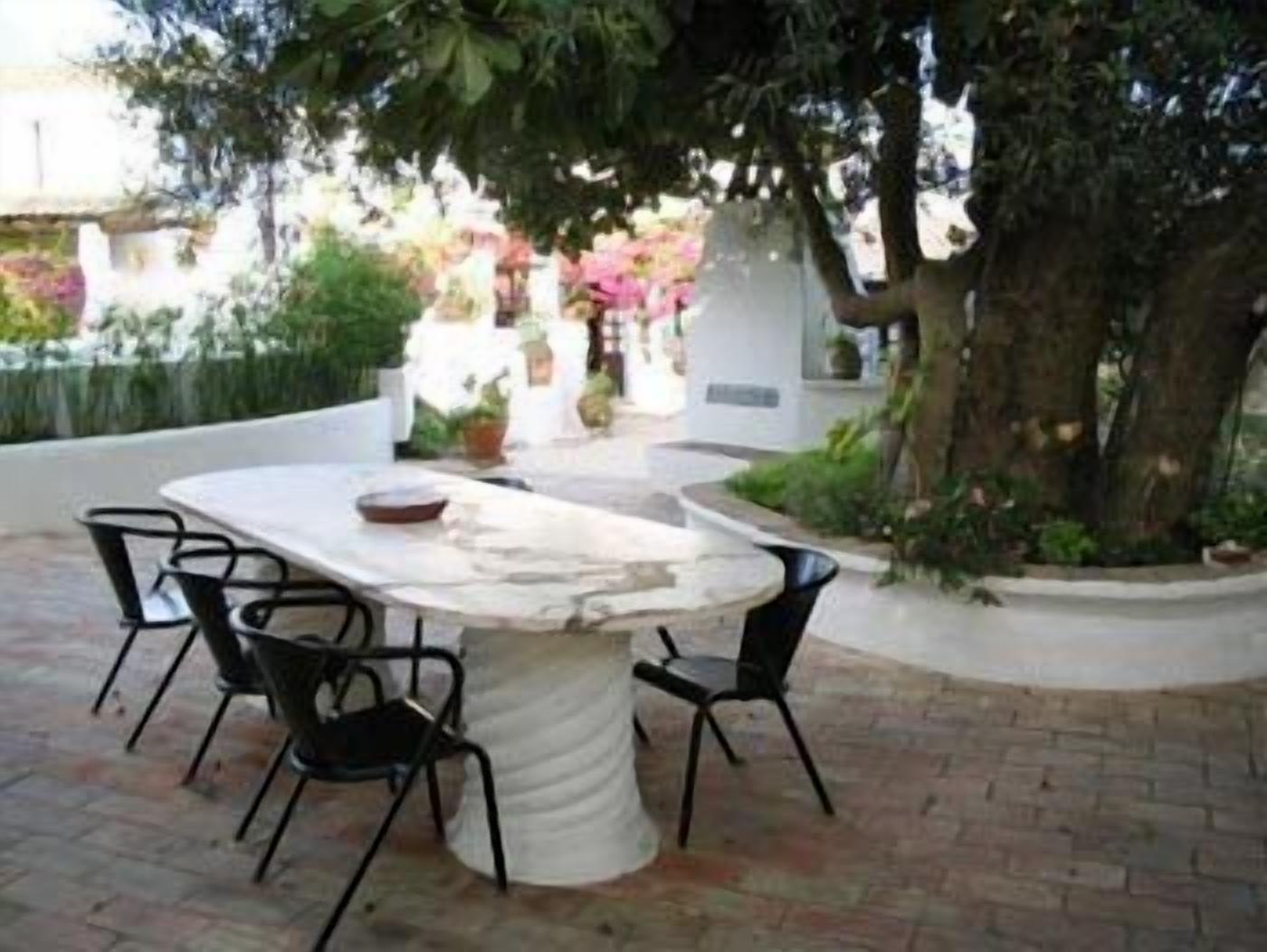 An outdoor patio with a marble table and four chairs, surrounded by greenery and a tree.