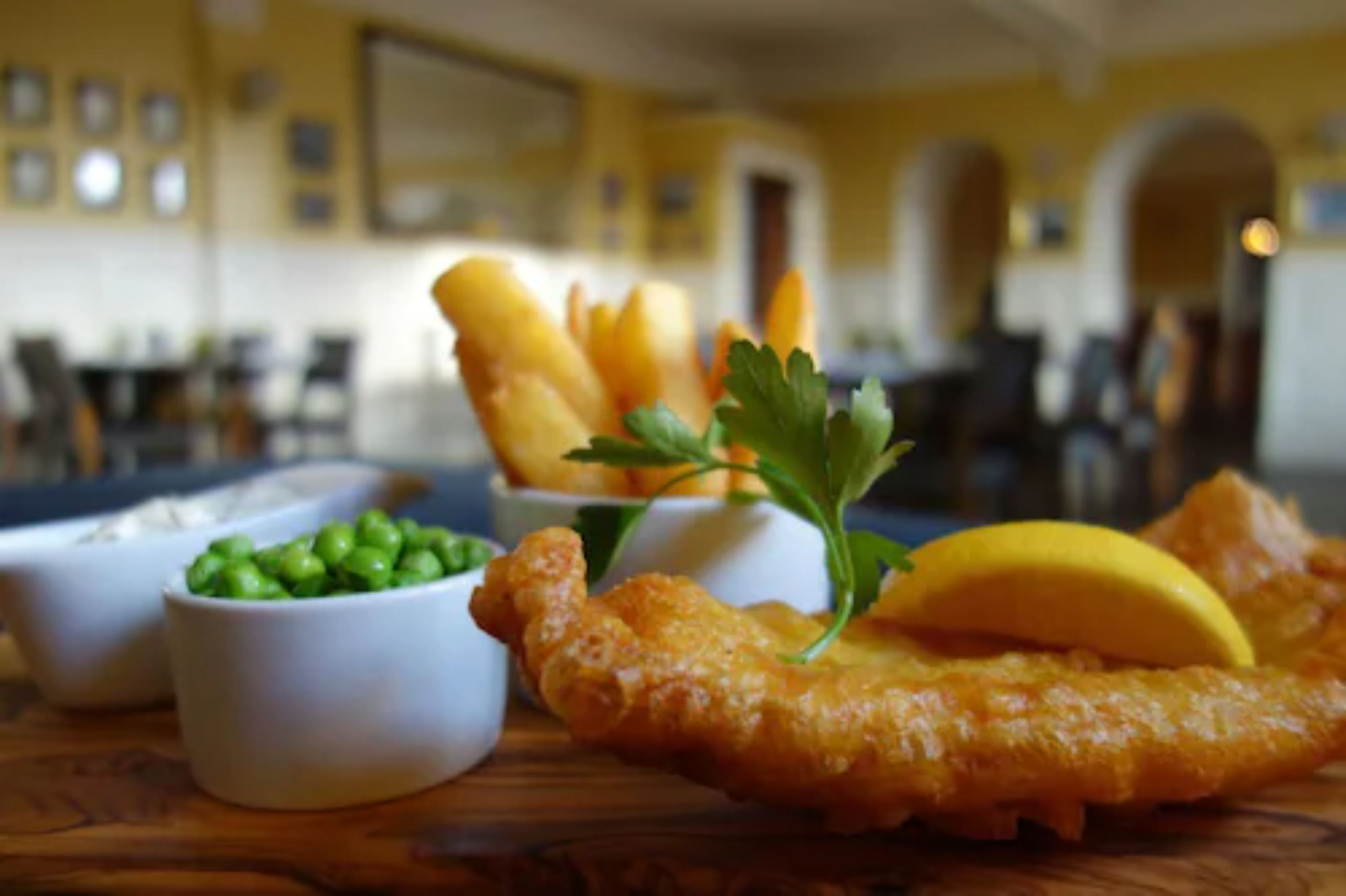 Una mesa con un plato grande de pescado frito, un gajo de limón y dos recipientes pequeños de guisantes y salsa tártara.