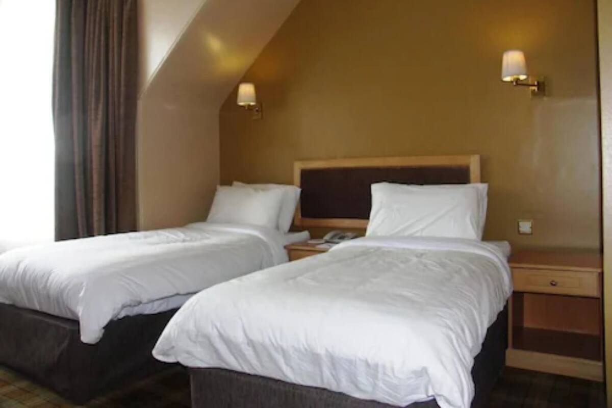A hotel room with two single beds, a wooden headboard, a nightstand with a lamp, and a window with curtains.