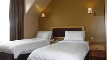 Habitación de hotel con dos camas individuales, cabecera de madera, mesita de noche con lámpara y ventana con cortinas.