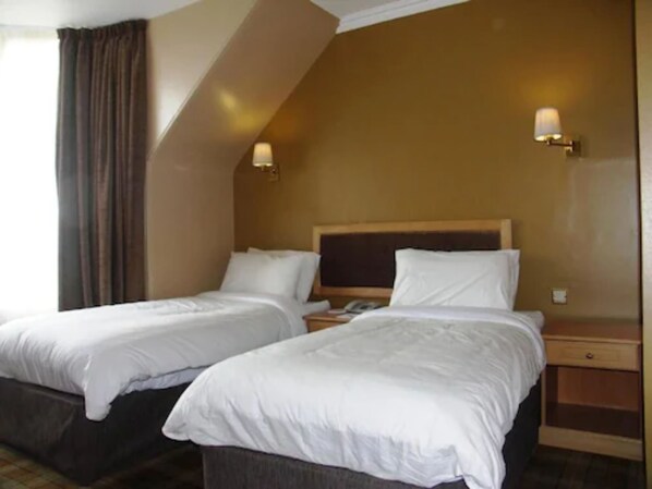 A hotel room with two single beds, a wooden headboard, a nightstand with a lamp, and a window with curtains. - Kilmarnock Arms Hotel (Peterhead)