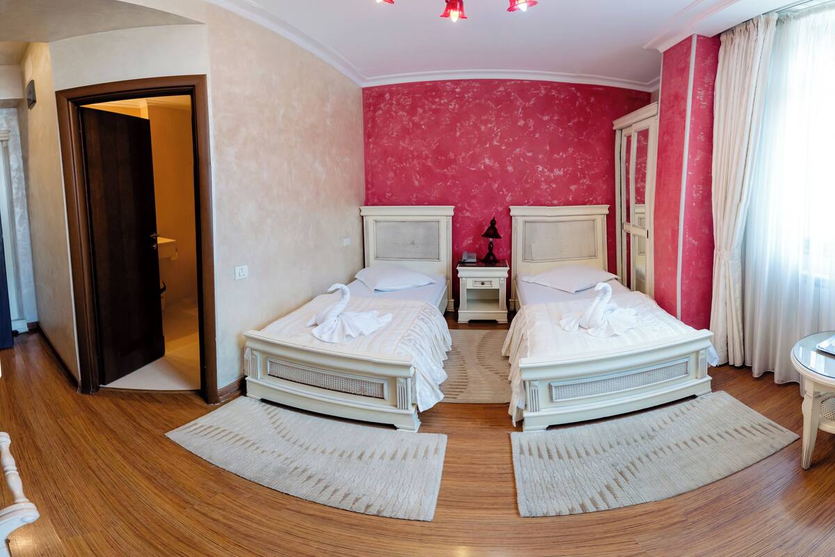 A hotel room with two beds, a wooden floor, a red accent wall, and a door leading to another room.