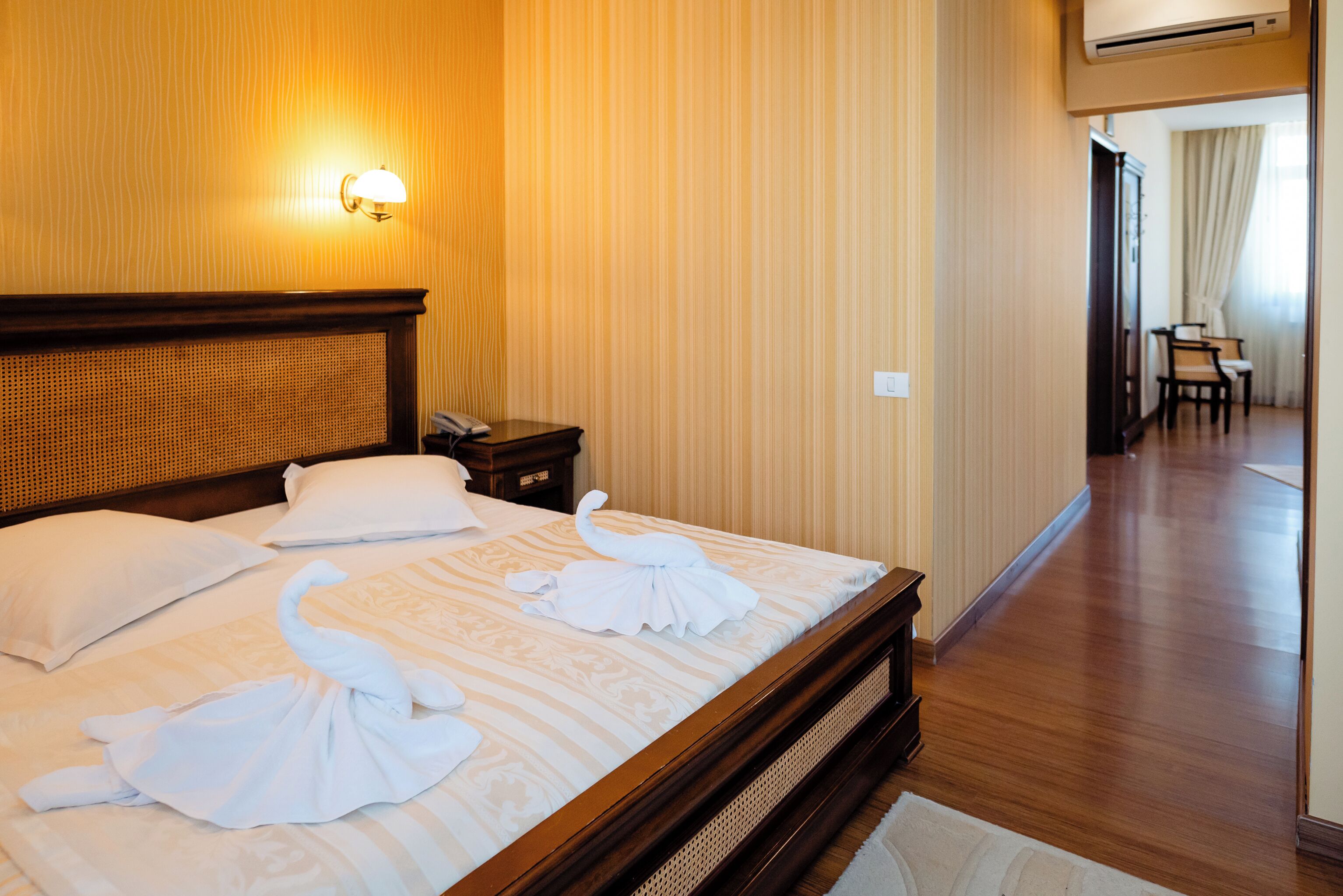 A hotel room with a wooden headboard, a bed with white linens and towels, a nightstand, and a wall-mounted lamp.