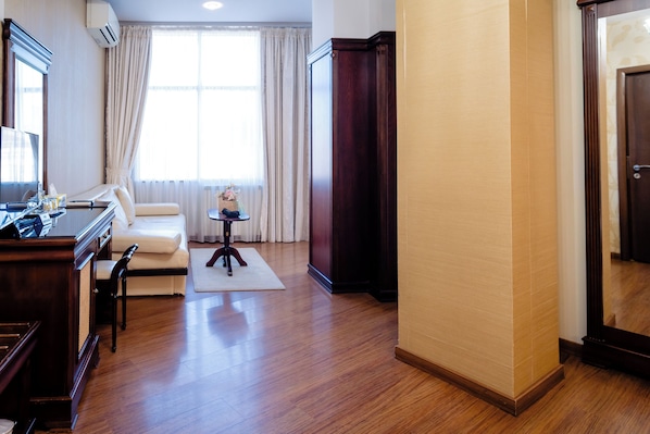 Una habitación de hotel con un escritorio de madera, un sofá, una mesita con un florero y un ventanal con cortinas.