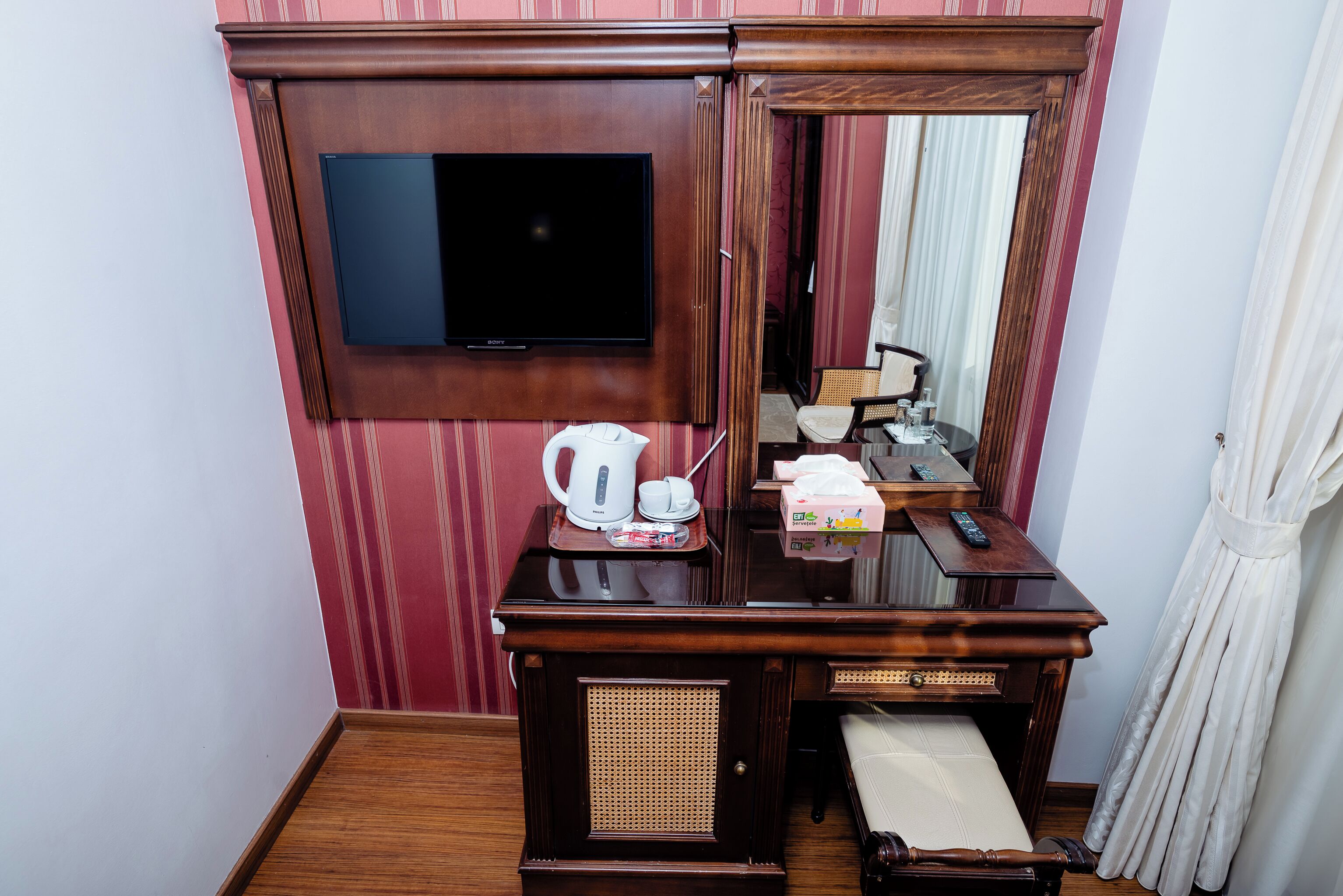 Habitació d'hotel amb escriptori de fusta, televisió penjada a la paret, mirall, cadira de vimetla i llit amb llençol blanc.