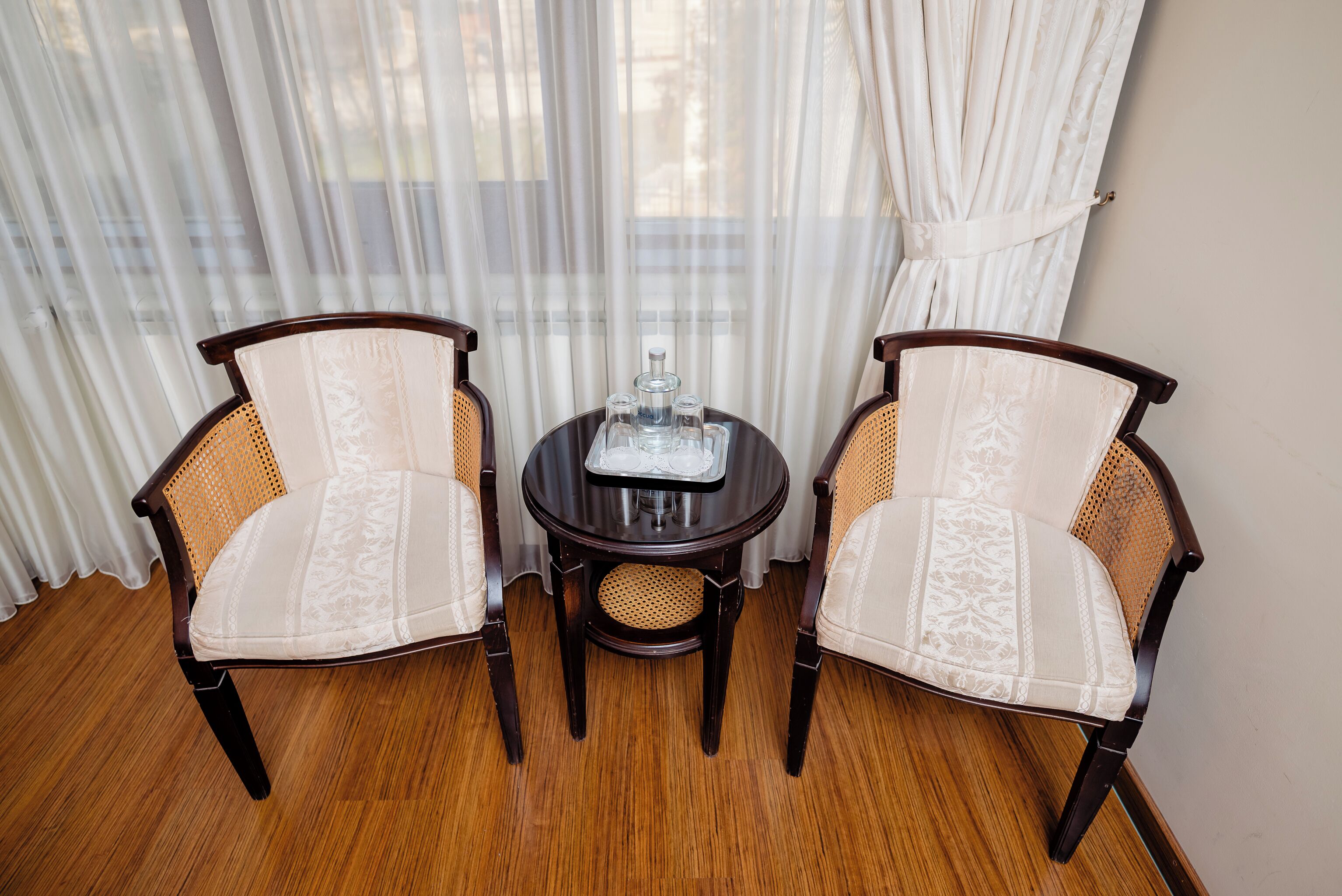 A room with two wooden chairs with woven backs and white cushioned seats, a small round table with a glass top, and a white curtain backdrop.
