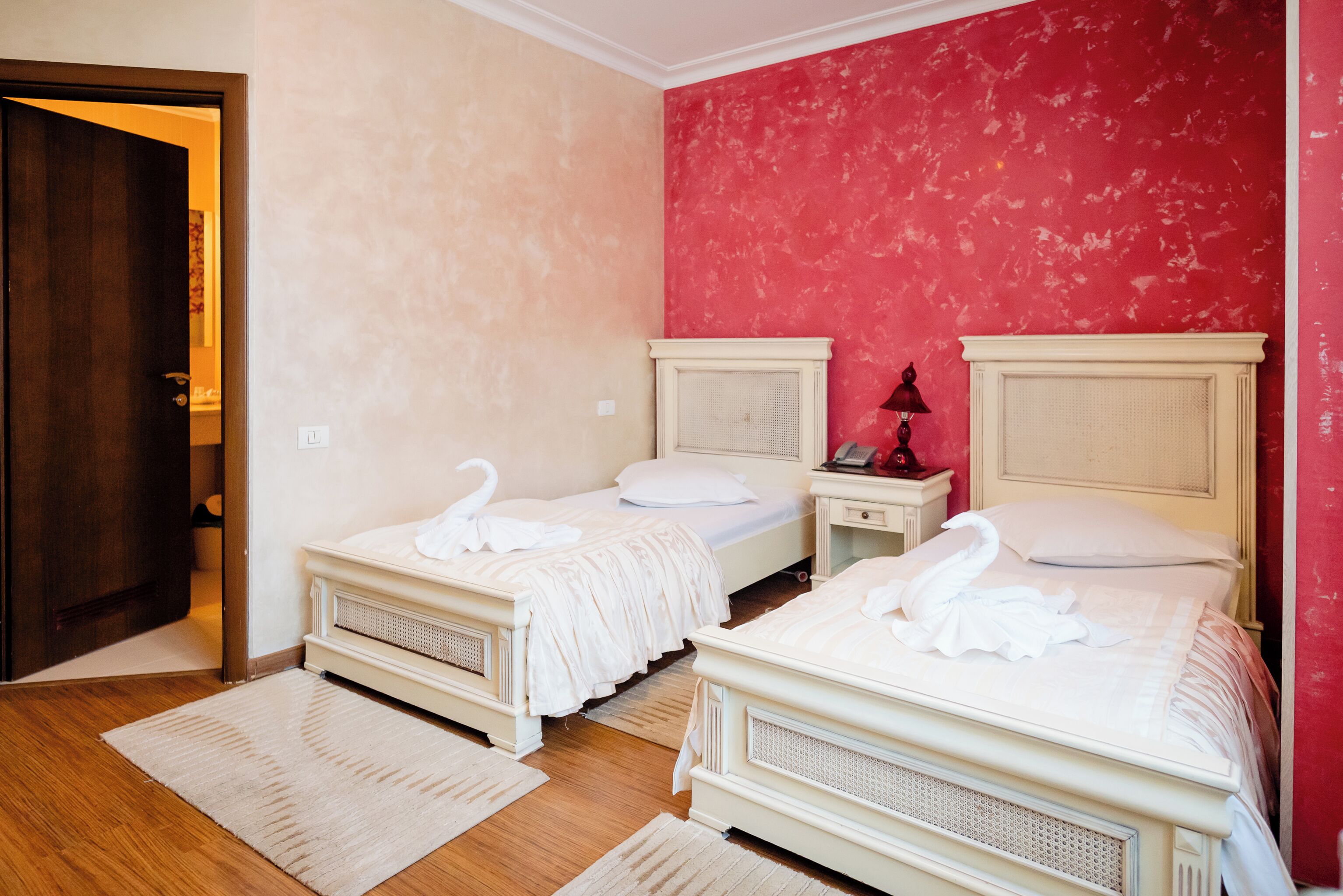 Habitación de hotel con dos camas, cada una con cabecero blanco y un cojín con forma de cisne. Hay una mesita de noche con una lámpara entre las camas, y una pared roja al fondo.