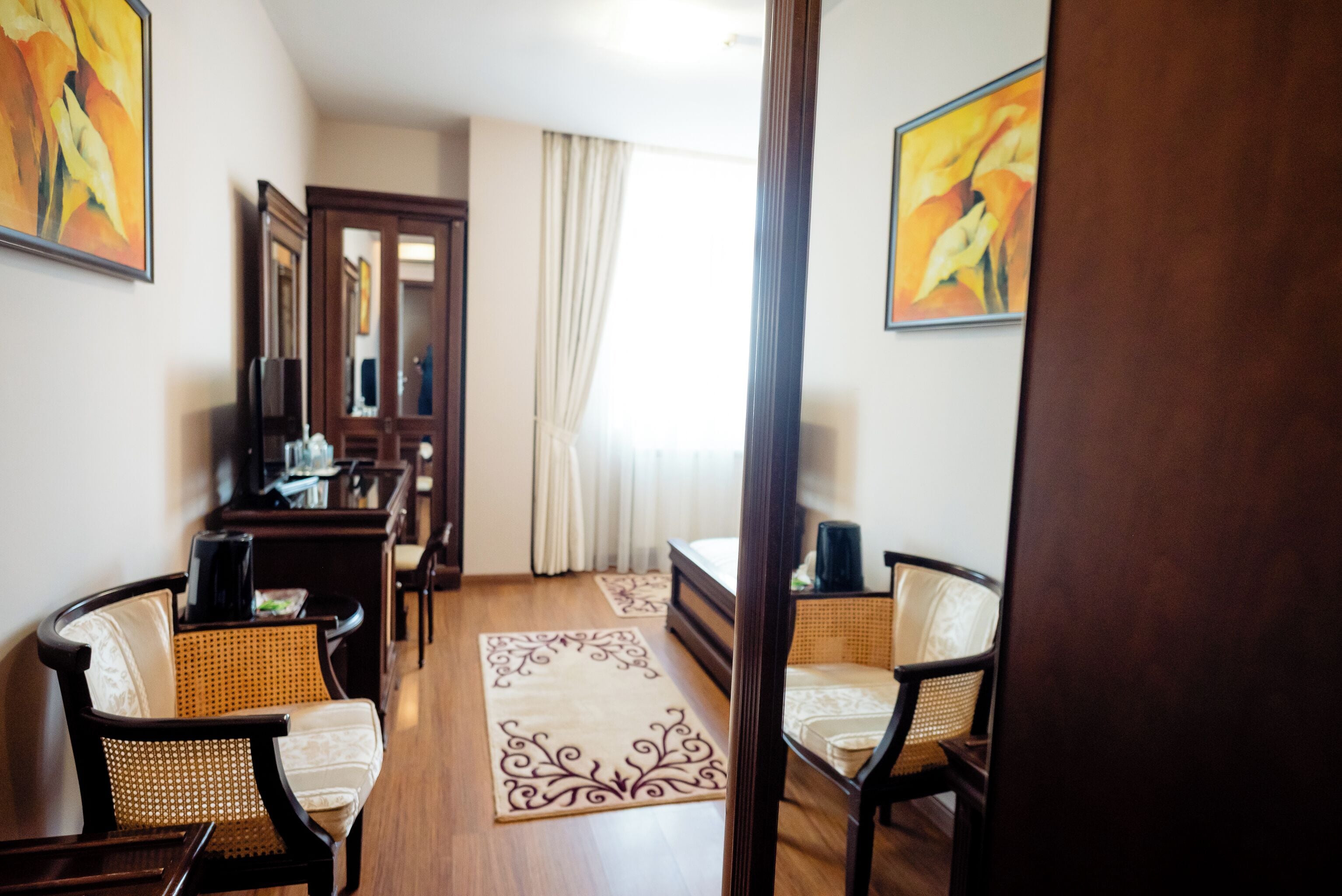A hotel room with a wooden floor, a bed with a white mattress, two wicker chairs, a desk with a computer, and a large mirror.