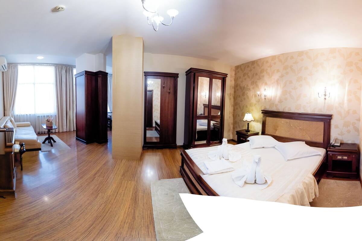 A hotel room with a wooden floor, a bed with white linens, a bedside table with a lamp, a wardrobe, and a mirror.